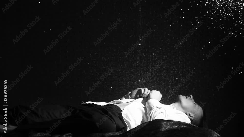 Man lies on a bed and buttoning a white shirt on black wall background at night. Loneliness, depression, fatigue and lack of motivation. Young man dreaming in bed before work