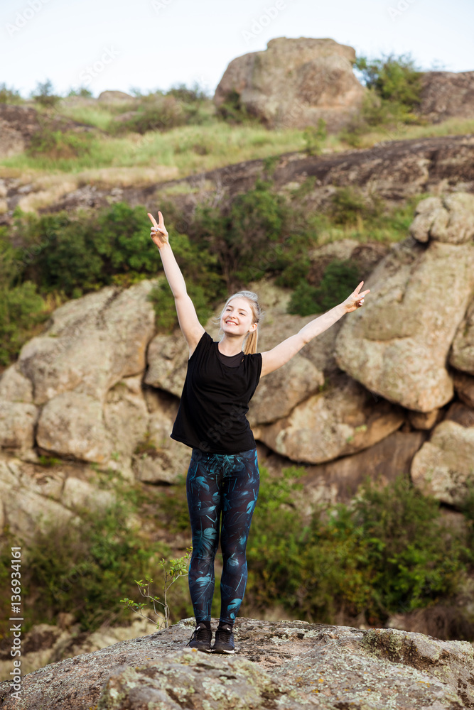 Sportive girl smiling, showing peace, standing on rock in canyon.