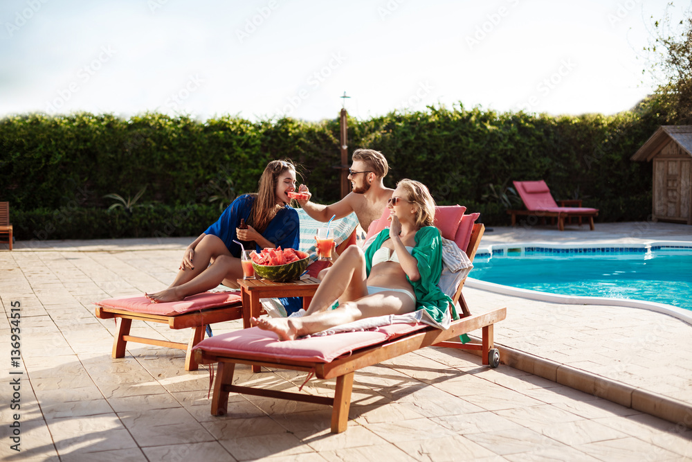 Friends smiling, eating watermelon, relaxing, lying near swimming pool.
