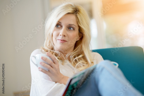 Middle-aged blond woman drinking coffee in armchair