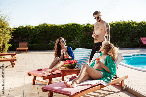 Friends smiling, sunbathing, drinking cocktails, lying near swimming pool.