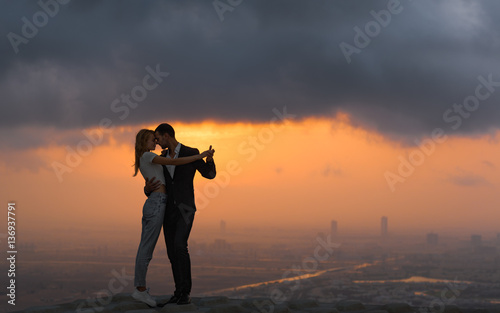 Two lovers dancing/embracing on top of a skyscraper overlooking the city at sunset. Romantic setting.