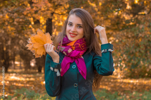 beautiful smiling girl in a pink scarf stands in the Park and keeps the leaves