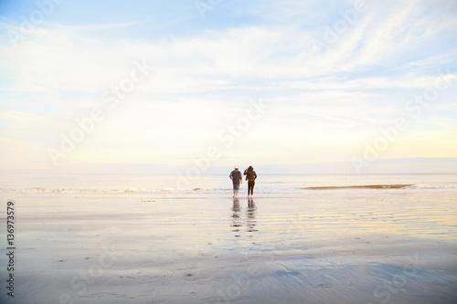 Couple standing on wet sand on beach and looking away. Man and woman relaxing on the sea ocean on sunset. Back view. Peaceful scene, calming waves, pastel cloudy sky, east coast. Happy time together.