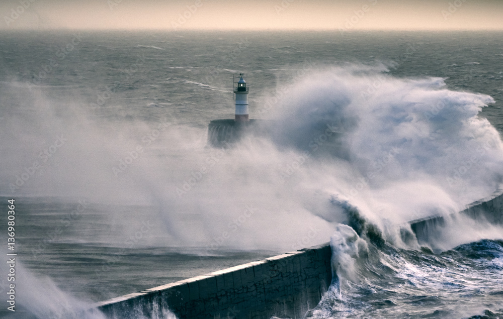 Huge waves crash over a sea wall near a lighthouse during a storm