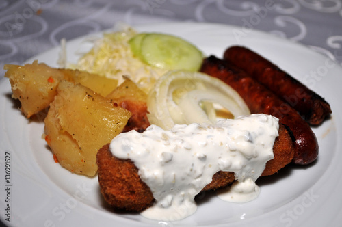 Beautifully decorated with the white plate of homemade dishes. Karađorđeva steak covered with a sauce of cream and leek, boiled potatoes, grilled sausages, fresh cucumbers and onions