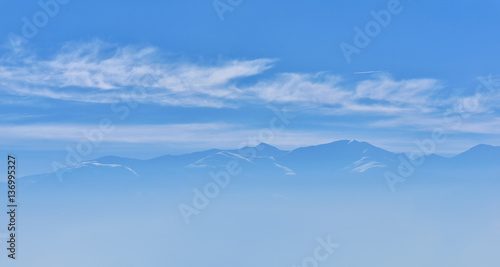 Beautiful view of mountain peak with sunrise light on top against blue sky with clouds in a foggy day © Daniel CHETRONI