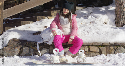 Pretty young asian woman in pink snowsuit relaxing on skiing holiday sat on wall. snowboard is laying next to her legs.
