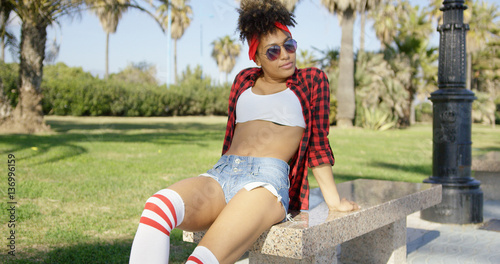 Sexy african american girl sitting on bench in tropical park. She relaxing on the sun. Wearing skimpy short jeans  checkered shirt and sunglasses.