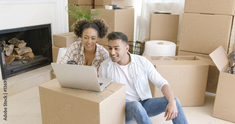 © Dash - Happy mixed race couple sitting on the floor among unpacked carton boxes and using laptop computer to plan the future in their new apartment they just moved in.