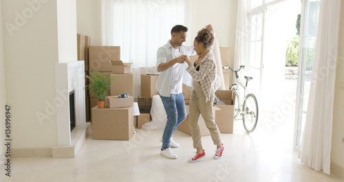 Happy young couple celebrating moving home dancing together in the living room surrounded by packing boxes