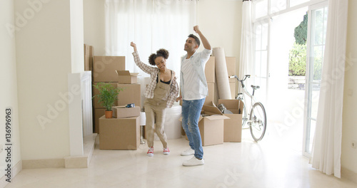 Young couple dancing for joy in the living room of their new home with packing boxes behind them