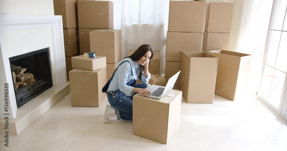Young woman using a laptop on a cardboard box as she catches up with her social media after moving into a new home Young woman using a laptop on a cardboard box as she catches up with her social media after moving into a new home