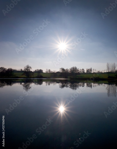 sun shining on a lake with a thin line of green fields in the middle, the sky, sun fields and trees are reflected in the lake duplicating the image in the lake photo