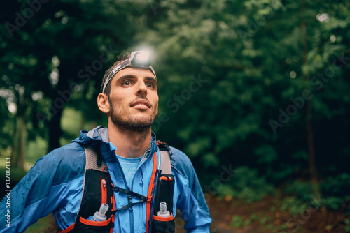Fit male jogger with a headlamp rests during training for cross country trail race in nature park.