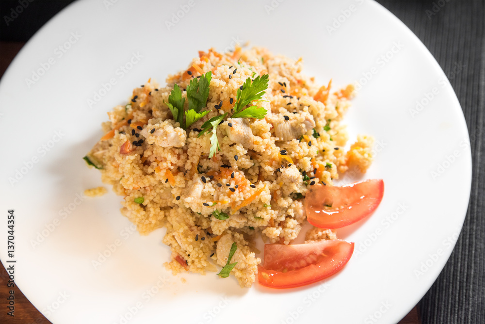 Couscous with grilled chicken, onions, carrots and tomatoes on a white plate on a dark background