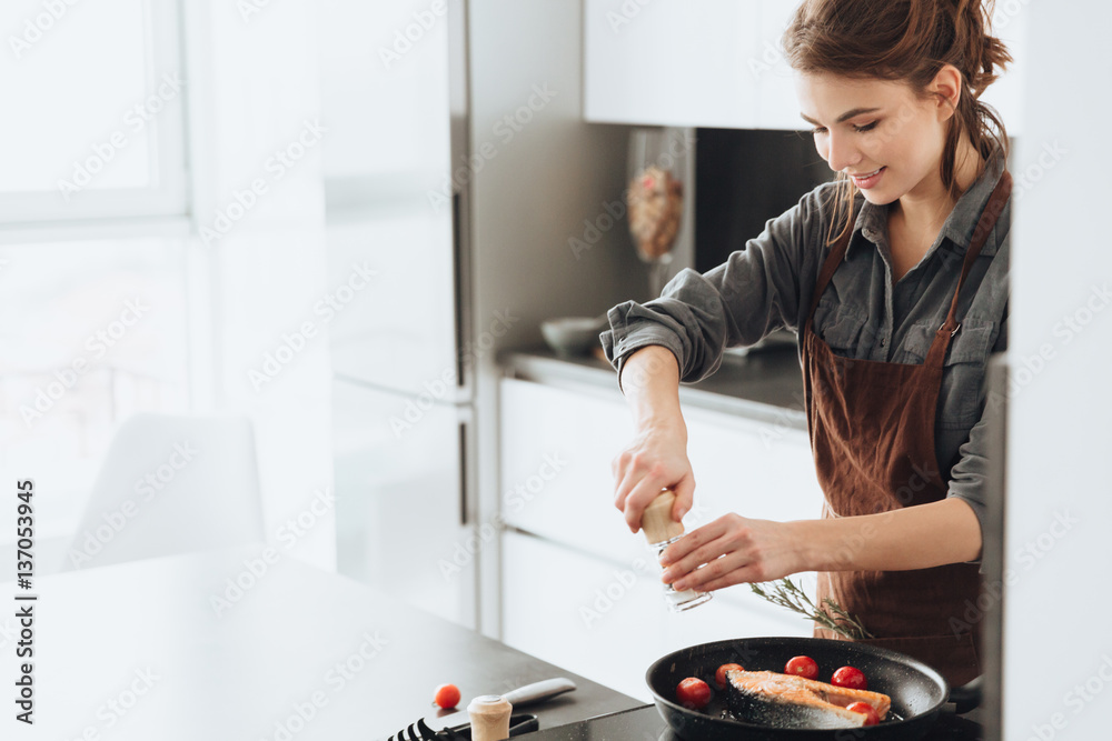 Pretty lady standing in kitchen while cooking fish - obrazy, fototapety, plakaty 