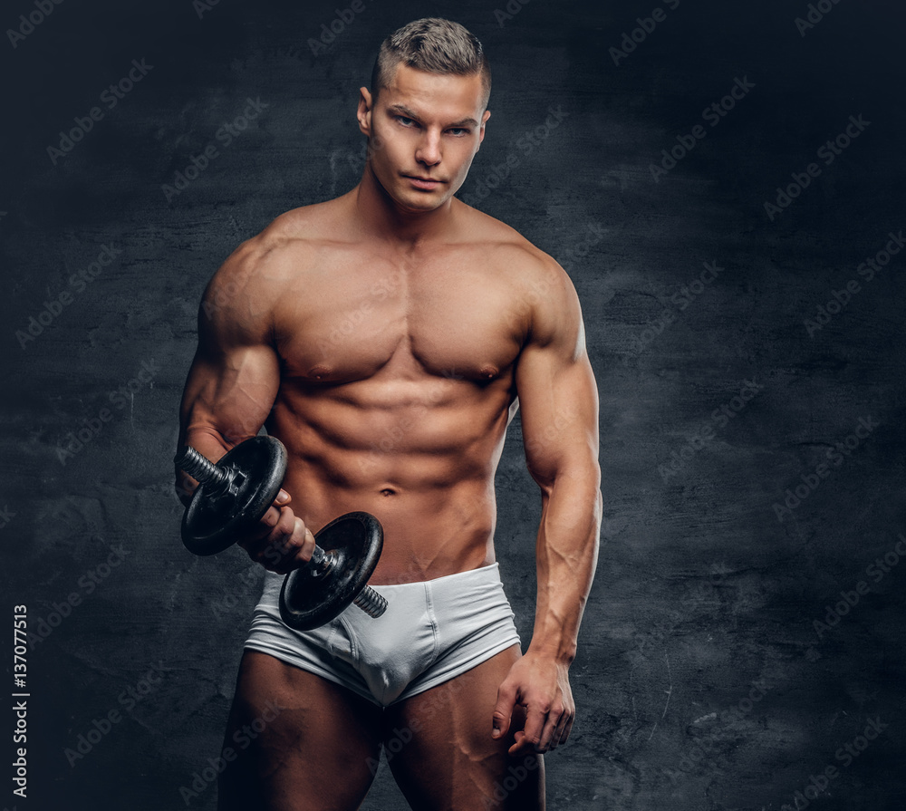 Shirtless muscular male holds dumbbell on grey background.
