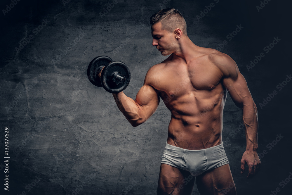 Shirtless muscular male holds dumbbell on grey background.