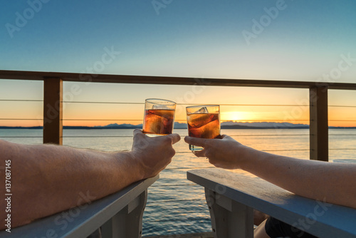 Couple holding up icy cocktail drinks while sitting on a seaside deck at sunset.