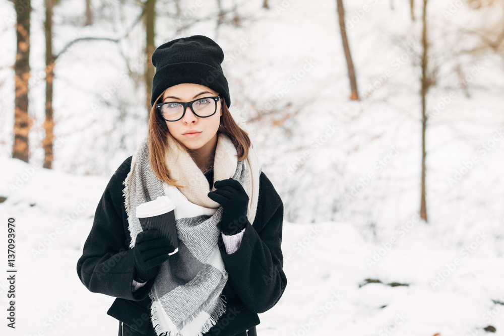 Portrait of a beautiful girl with blue eyes in black glasses in a black hat and coat and gloves with scarf in the winter park holding warm coffee Portrait of a beautiful girl with blue eyes in black glasses in a black hat and coat and gloves with scarf in the winter park holding warm coffee