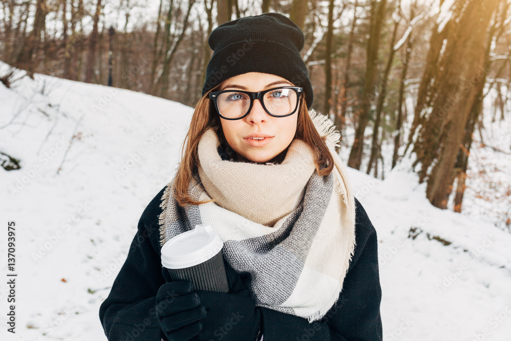 Portrait of a beautiful girl in black glasses in a black hat and coat and gloves with scarf in the winter park holding warm coffee Portrait of a beautiful girl in black glasses in a black hat and coat and gloves with scarf in the winter park holding warm coffee