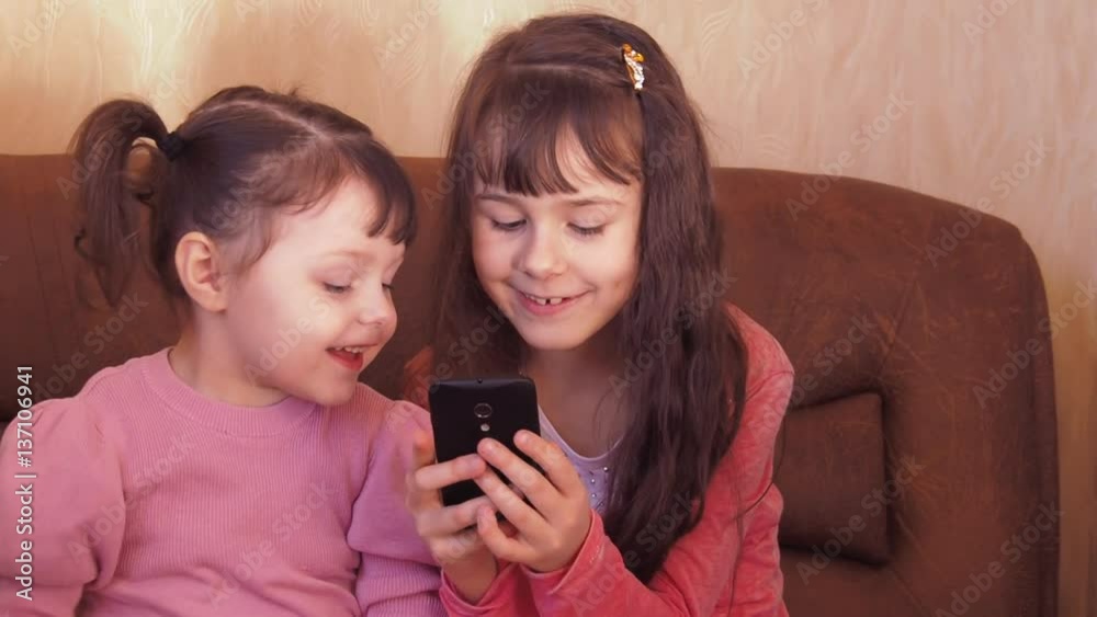 Little girls with a mobile phone. Two little girls looking at mobile phone smart phone at home on the couch Full hd Little girls with a mobile phone. Two little girls looking at mobile phone smart phone at home on the couch Full hd