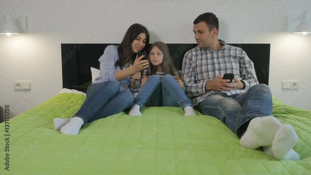 Young woman, man and their little daughter using gadgets on big bed in bedroom