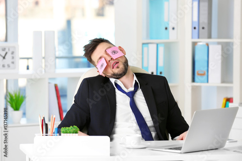 Schilderij op canvas Young business man with fake eyes painted on paper stickers sleeping at workplac