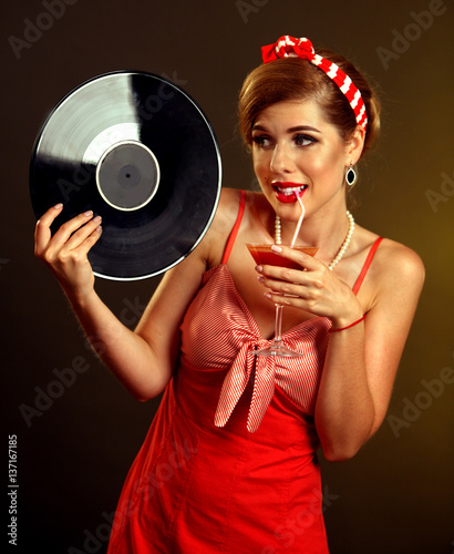 Retro woman with music vinyl record. Pin up girl drink martini cocktail . Pin-up retro female style. Girl wearing red dress drinking beverage through straw and is lost in thought.