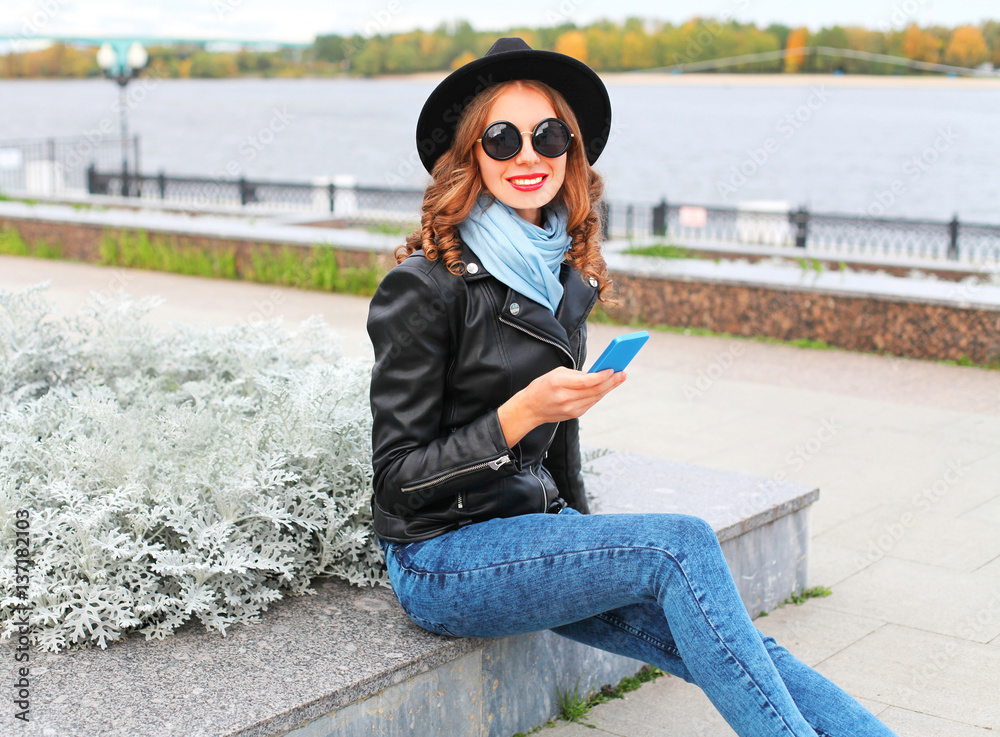 Fototapeta premium Fashion young smiling woman using smartphone wearing black rock jacket, hat in city park