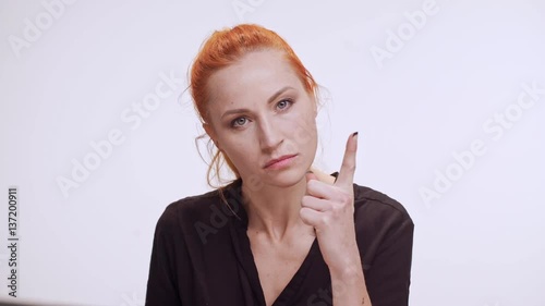 Strict middle-aged Caucasian woman with colored orange hair pointing finger standing on white background in dark brown shirt
