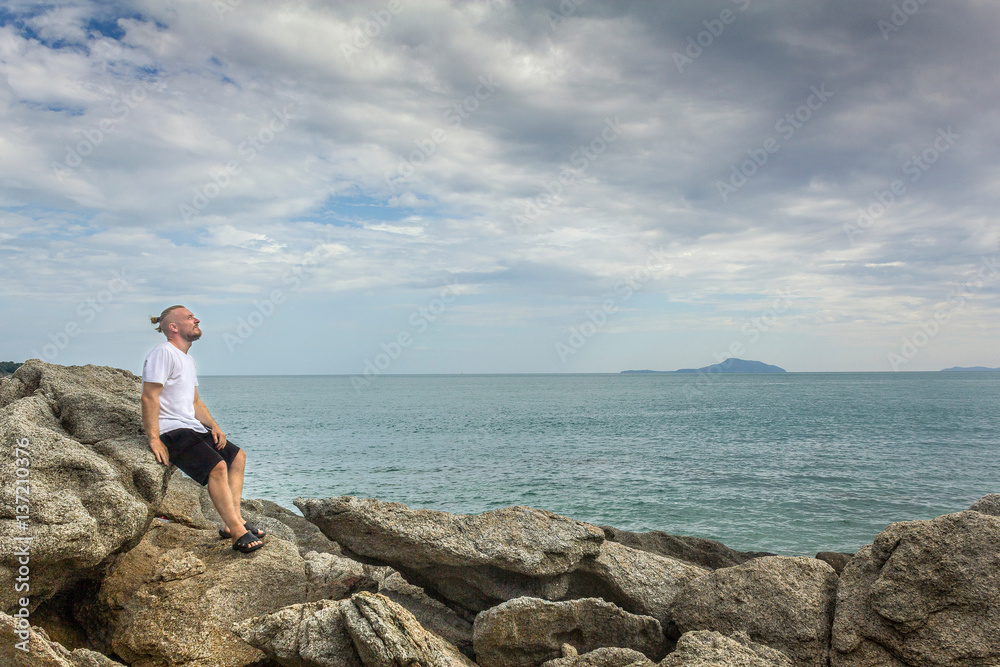 Man looking at sky near sea. Side view of man admiring cloudscape sitting on stones by the ocean. 