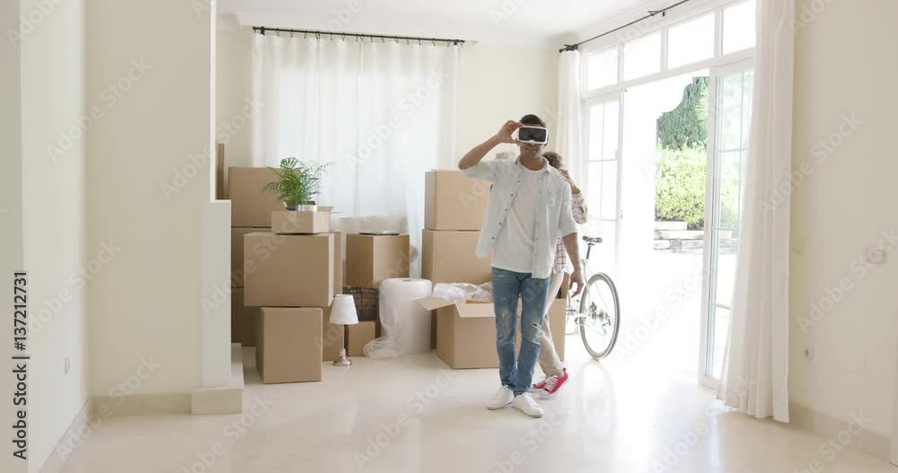 Young man trying out virtual reality goggles guided by his wife standing in a bare living room surrounded by brown boxes during a move to a new home. Young man trying out virtual reality goggles guided by his wife standing in a bare living room surrounded by brown boxes during a move to a new home.