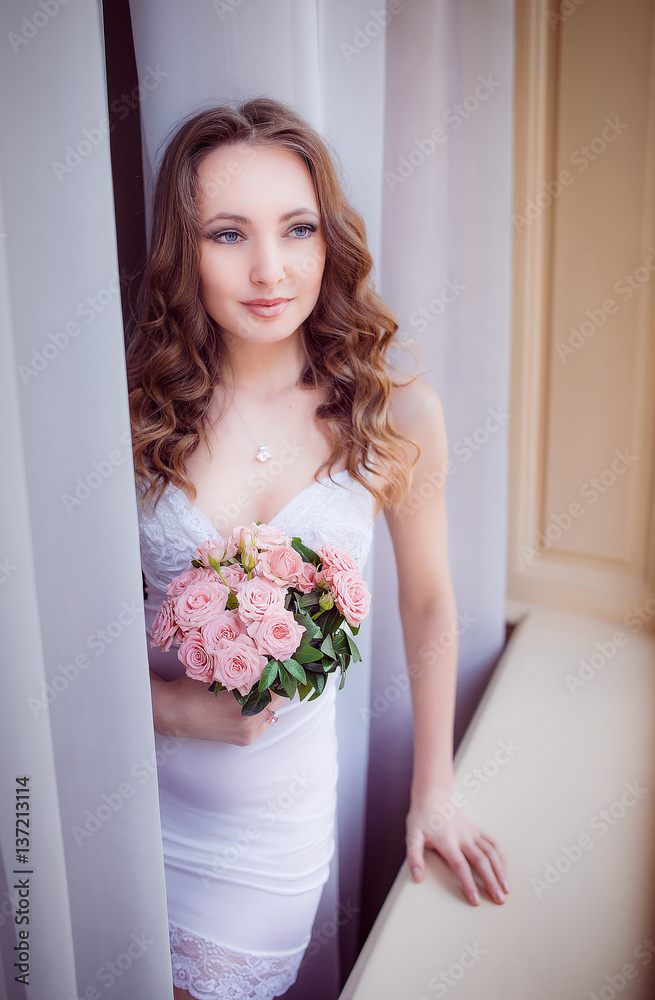 Beautiful bride stands with wedding bouquet before windowsill