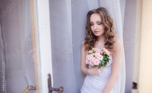 Elegant bride in white lingerie holds pink bouquet standing before the window