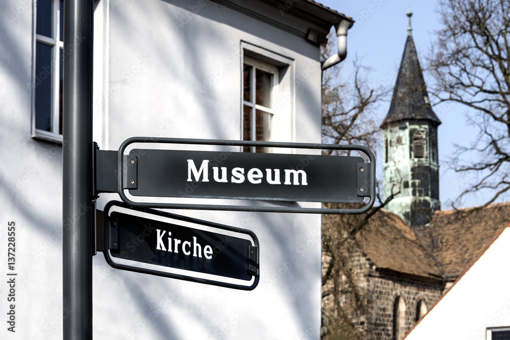 Fototapeta premium German signs indicate direction to Museum and Church with houses and steeple of a church in the background.