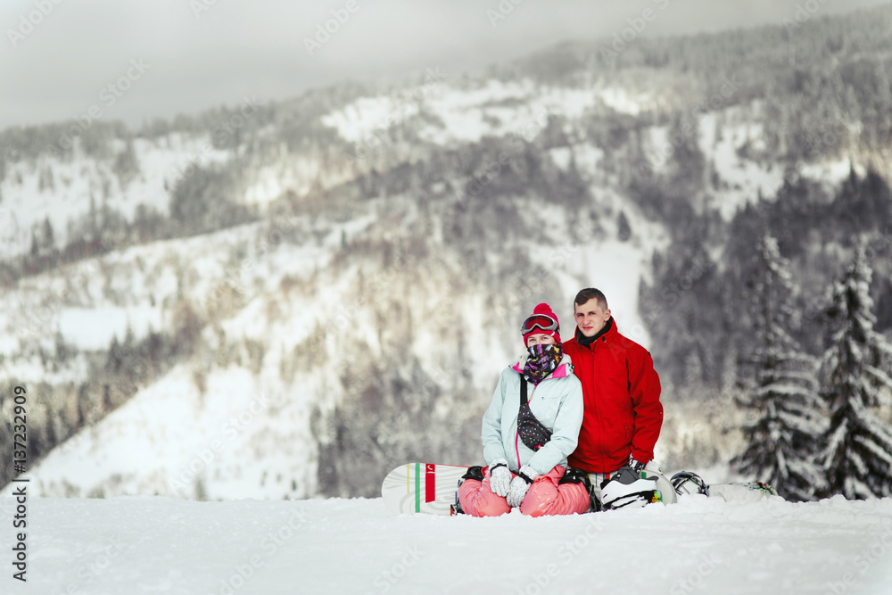 Man in red ski jacket lies his hand on woman's shoulder while they pose on snowed hill in the mountains