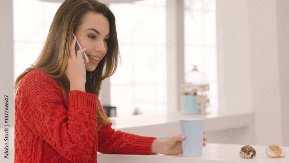 Side view of Smiling Woman in red sweater sitting by the table in cafe, talking on smartphone and drinking coffee Side view of Smiling Woman in red sweater sitting by the table in cafe, talking on smartphone and drinking coffee