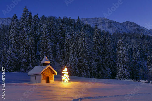 Leinwand Poster Illuminated Christmas tree in front of a chapel in winter, Bavaria, Upper Bavari