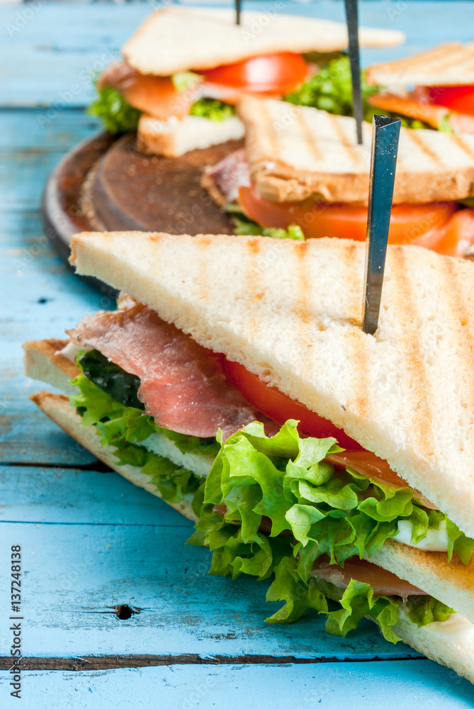 Obraz premium Club sandwiches with fresh lettuce, tomato, cucumber and salmon (trout). On crunchy toast. On blue rustic table, picnic, close view copy space