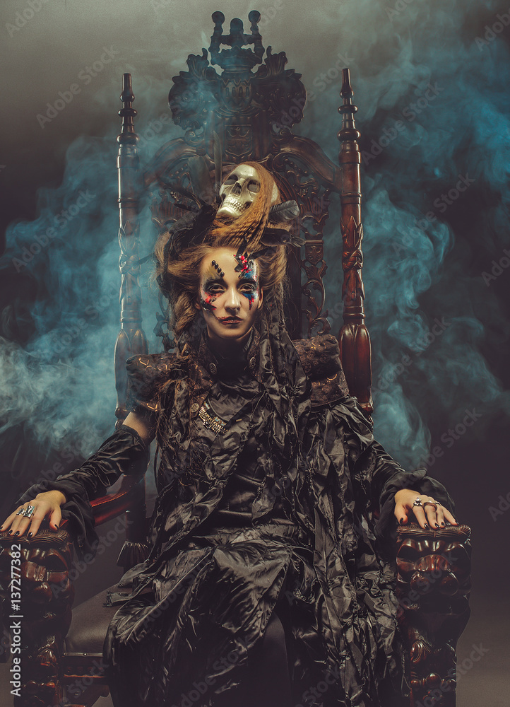 Young beautiful witch sits on a chair. Bright make up, skull, smoke-  halloween theme.