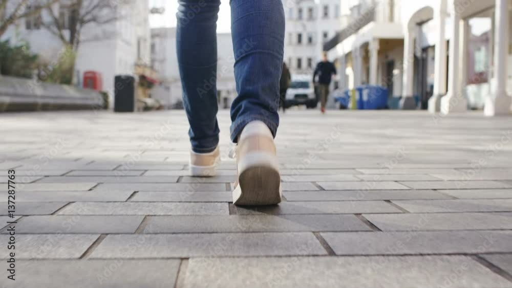 4K Camera follows behind the feet of a female walking through town, in slow motion