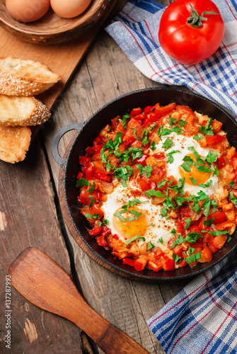 shakshuka - fried eggs, onion, bell pepper, tomatoes and parsley in a pan on a rustic wooden table with ingredients. top view photo