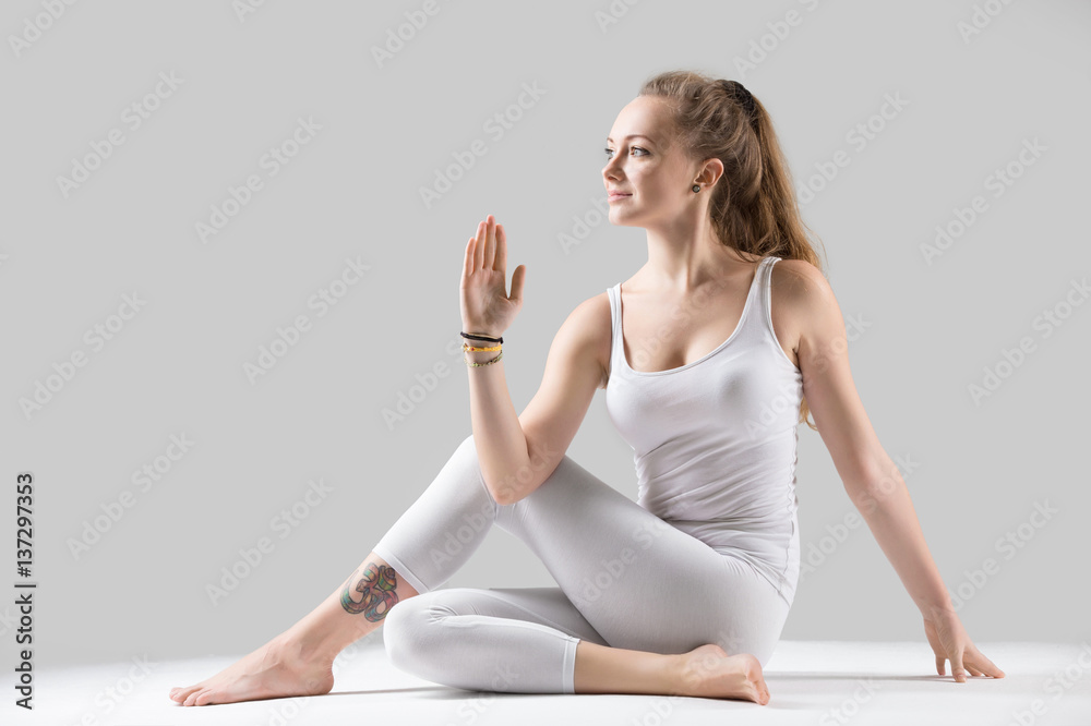 Young attractive woman practicing yoga, sitting in Ardha Matsyendrasana exercise, Half lord of the fishes pose, working out, wearing sportswear, white tank top, pants, indoor full length, grey studio