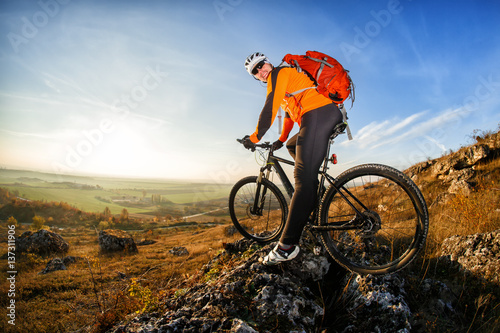 Cyclist in helmet and glasses on mountain bike stands on the precipice of hill under blue sky and sun. Wide angle view