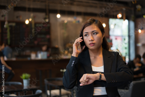 Beautiful business woman talking on cell phone looking at hand watch over cafe background.