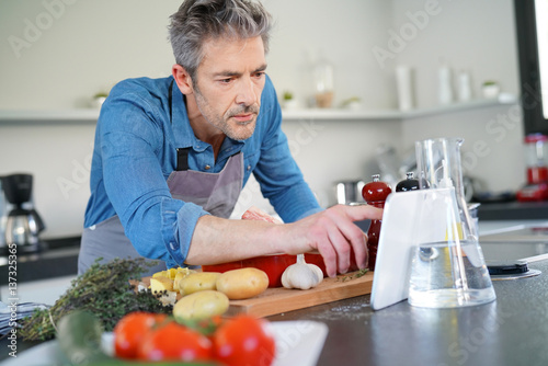 Quadro em tela Mature man in kitchen cooking with help of receipe on tablet