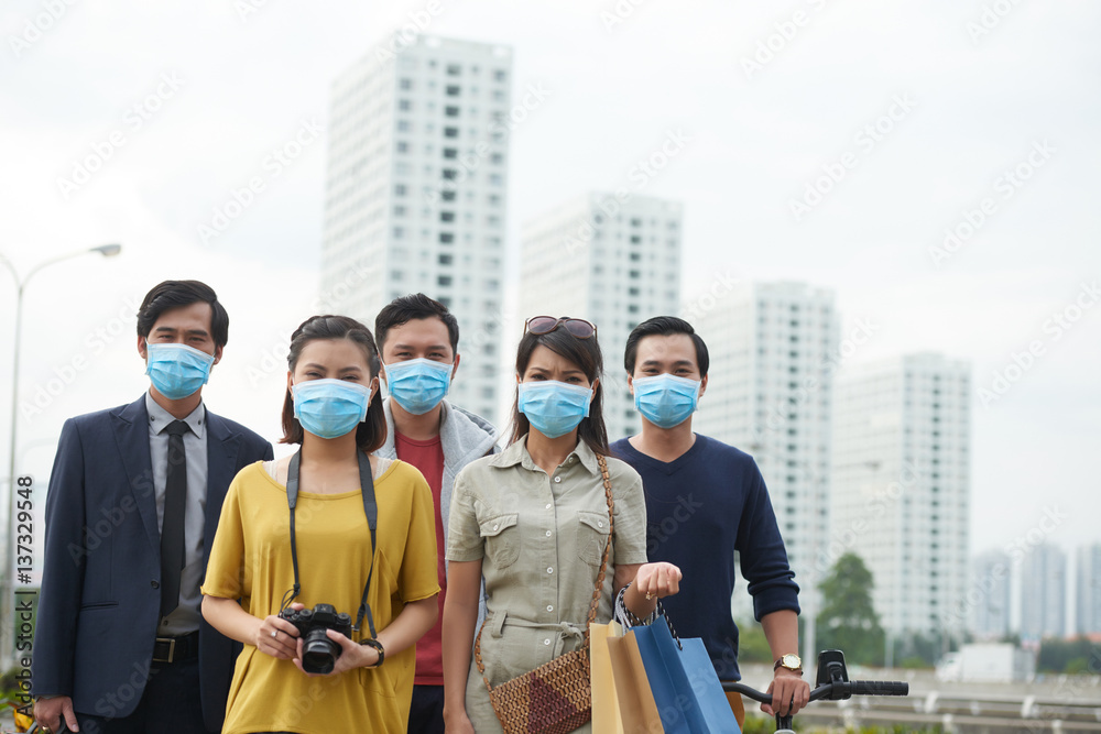 Waist-up portrait of Asian friends in protective masks looking at camera while standing against modern skyscrapers