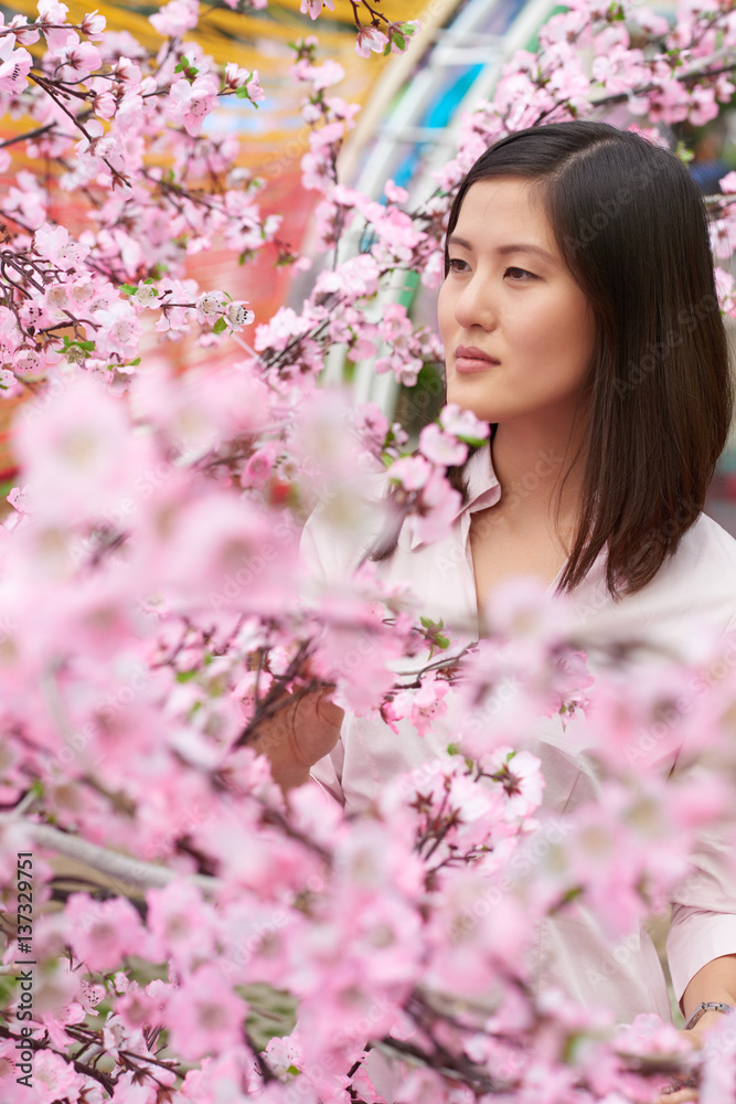 Thoughtful Asian woman standing at blossoming cherry tree and enjoying its beauty, waist-up portrait